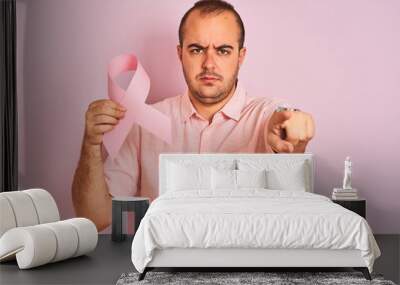 Young man holding cancer ribbon symbol standing over isolated pink background pointing with finger to the camera and to you, hand sign, positive and confident gesture from the front Wall mural