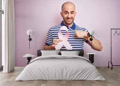 Young man holding pink brest cancer ribbon over isolated background with surprise face pointing finger to himself Wall mural