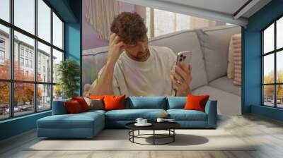 Young man sitting on a couch at home looking at his smartphone in a relaxed manner within a cozy living room interior featuring soft furnishings Wall mural