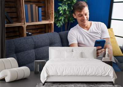 Young man using smartphone sitting on sofa at home Wall mural