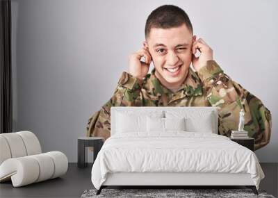 Young man wearing camouflage army uniform covering ears with fingers with annoyed expression for the noise of loud music. deaf concept. Wall mural