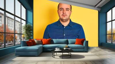 Young man wearing denim casual shirt standing over isolated yellow background with serious expression on face. Simple and natural looking at the camera. Wall mural