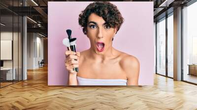 Young man wearing woman make up holding set of brushes scared and amazed with open mouth for surprise, disbelief face Wall mural