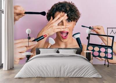 Young man wearing woman make up with stylist all around him peeking in shock covering face and eyes with hand, looking through fingers afraid Wall mural