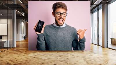 Young man with beard wearing glasses holding broken and craked smartphone pointing and showing with thumb up to the side with happy face smiling Wall mural