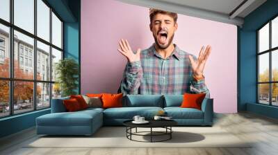 Young man with beard wearing golden crown of king standing over isolated pink background celebrating mad and crazy for success with arms raised and closed eyes screaming excited. Winner concept Wall mural