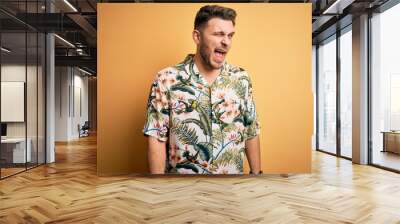 Young man with blue eyes on vacation wearing floral summer shirt over yellow background winking looking at the camera with sexy expression, cheerful and happy face. Wall mural