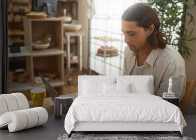 Young man with long hair using laptop and smartphone inside a cozy bakery shop, surrounded by shelves filled with bread and pastries. Wall mural