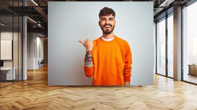 Young man with tattoo wearing orange sweater standing over isolated white background smiling with happy face looking and pointing to the side with thumb up. Wall mural