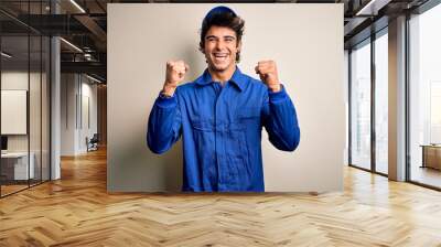 Young mechanic man wearing blue cap and uniform standing over isolated white background celebrating surprised and amazed for success with arms raised and open eyes. Winner concept. Wall mural