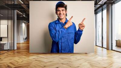 Young mechanic man wearing blue cap and uniform standing over isolated white background smiling and looking at the camera pointing with two hands and fingers to the side. Wall mural