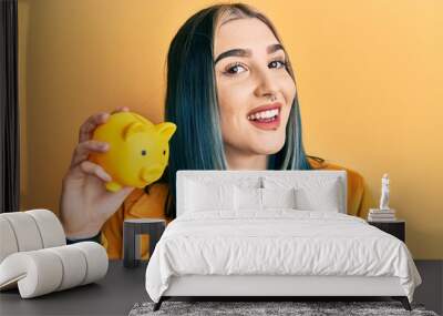 Young modern girl holding yellow piggy bank looking positive and happy standing and smiling with a confident smile showing teeth Wall mural