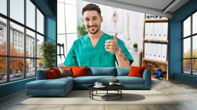 Young physiotherapist man working at pain recovery clinic doing happy thumbs up gesture with hand. approving expression looking at the camera showing success. Wall mural