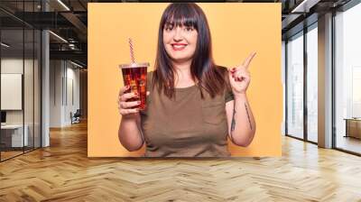 Young plus size woman drinking glass of cola beverage smiling happy pointing with hand and finger to the side Wall mural