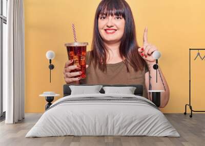 Young plus size woman drinking glass of cola beverage smiling with an idea or question pointing finger with happy face, number one Wall mural