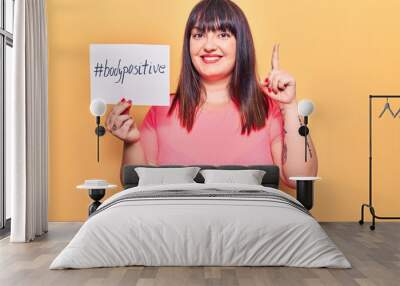 Young plus size woman holding paper with hashtag body positive smiling with an idea or question pointing finger with happy face, number one Wall mural