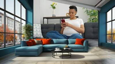 Young pregnant woman using smartphone sitting on sofa at home Wall mural