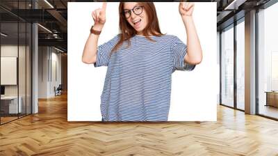 Young read head woman wearing casual clothes and glasses smiling amazed and surprised and pointing up with fingers and raised arms. Wall mural