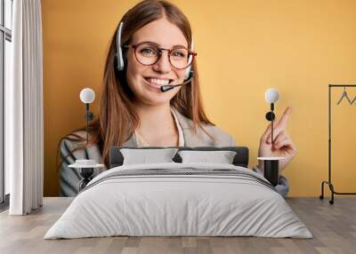 Young redhead call center agent woman overworked wearing glasses using headset with a big smile on face, pointing with hand and finger to the side looking at the camera. Wall mural