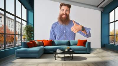 Young redhead irish man wearing casual shirt standing over isolated white background doing happy thumbs up gesture with hand. Approving expression looking at the camera with showing success. Wall mural