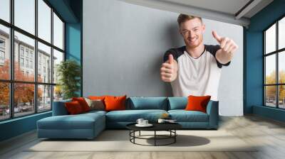 Young redhead man over grey grunge wall approving doing positive gesture with hand, thumbs up smiling and happy for success. Looking at the camera, winner gesture. Wall mural