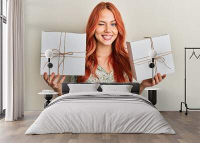 Young redhead woman holding gifts winking looking at the camera with sexy expression, cheerful and happy face. Wall mural