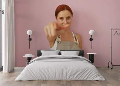 Young redhead woman pointing at camera over isolated pink background wearing an apron. Wall mural