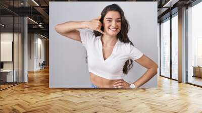 Young teenager girl standing over white background smiling doing phone gesture with hand and fingers like talking on the telephone. communicating concepts. Wall mural