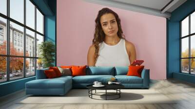 Young woman holding a credit card and smartphone against an isolated pink background, expressing thoughtful contemplation, with attractive hispanic brunette features in casual attire Wall mural