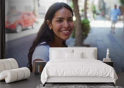 Young woman holding a phone and smiling on a city street, exuding happiness and vibrancy in an urban setting. Wall mural