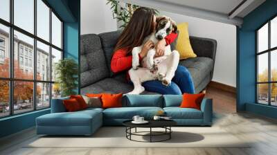 Young woman kissing and hugging dog sitting on sofa at home Wall mural