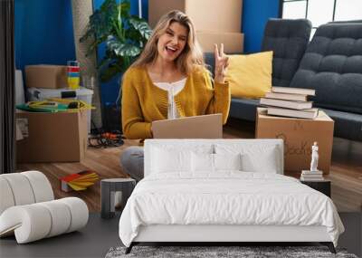 Young woman sitting on the floor at new home using laptop smiling with happy face winking at the camera doing victory sign. number two. Wall mural