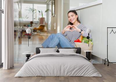 Young woman sitting on the kitchen floor with a paper bag full of fresh groceries smiling in love showing heart symbol and shape with hands. Romantic concept. Wall mural