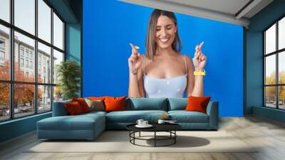 Young woman standing over blue background gesturing finger crossed smiling with hope and eyes closed. luck and superstitious concept. Wall mural