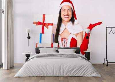 Young woman wearing santa claus costume holding gifts celebrating victory with happy smile and winner expression with raised hands Wall mural