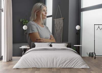 Young woman with blonde hair standing indoors in a modern living room, looking thoughtfully out the window with arms crossed Wall mural