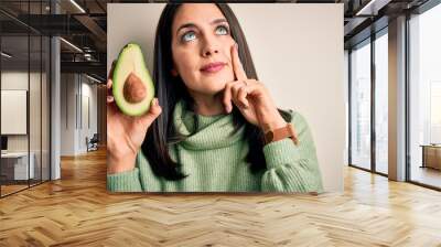 Young woman with blue eyes holding middle healthy avocado over isolated white background serious face thinking about question, very confused idea Wall mural