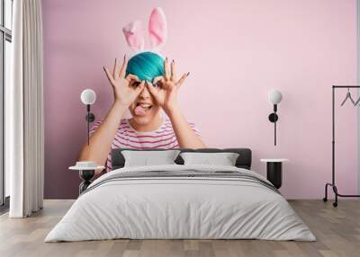 Young woman with fashion blue hair wearing easter rabbit ears over pink background doing ok gesture like binoculars sticking tongue out, eyes looking through fingers. Crazy expression. Wall mural