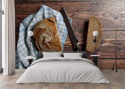 A round loaf of a crusty bread, on a rustic wooden table. With a bread knife and a blue and white checkered kitchen towel. Wall mural