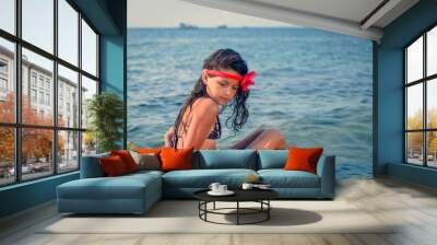 Mysterious dreamy young girl with a red bandage on her head sitting in the water on a rocky beach Wall mural