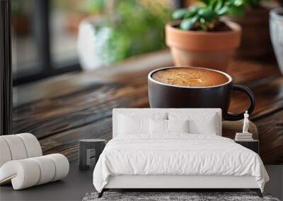 A cup of coffee on a wooden table, with a plant and pot blurred in the background Wall mural