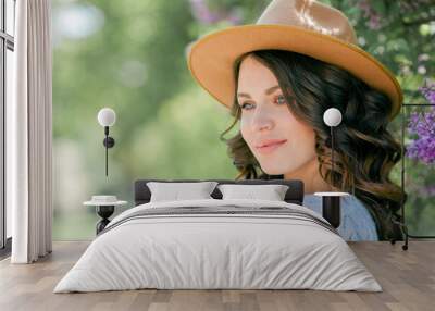 Portrait of beautiful young dark-haired woman with blue eyes in hat near the blooming. Happy model with curly hair. Spring. Wall mural