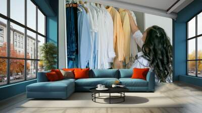 Young dark-haired curly beautiful woman is sorting things in large spacious closet. Home organization. Washing things. Analysis of clothes by season. Wall mural