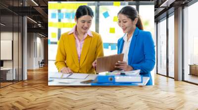 Collaborative Brainstorming: Two professional women, one in a yellow blazer and the other in a blue blazer, engage in a lively discussion, reviewing ideas on a clipboard. Wall mural