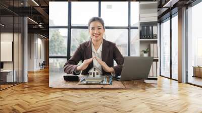 Real Estate Agent Protecting Dream Home: A confident and friendly Asian woman real estate agent in a professional suit, sitting at her desk, makes a protective gesture over a miniature house model. Wall mural