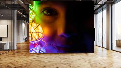 portrait of a little seven year old kid girl holding a garland in her hands near her face, glowing with multicolored neon lights at night time at home. Christmas Eve holiday celebration. banner Wall mural