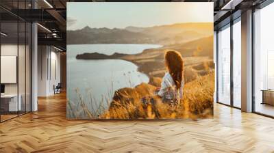 A beautiful young woman tourist is sitting on top of a mountain and enjoying the view of the sunset sea and mountains. The concept of freedom and meditation, an active lifestyle. Wall mural