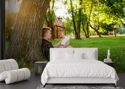 A young man is sitting under a tree and reading a book. Green summer park at sunset Wall mural