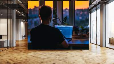 A young man programmer coding on a laptop in the dark with a view of the lights of the night city, color lighting in the room. Coding and hacking concept Wall mural