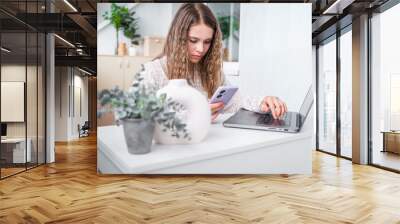 A young teenage student girl looking a laptop screen, online courses, studying remotely from home, sitting at a desk with a phone and laptop in a modern bright interior with plants. Wall mural